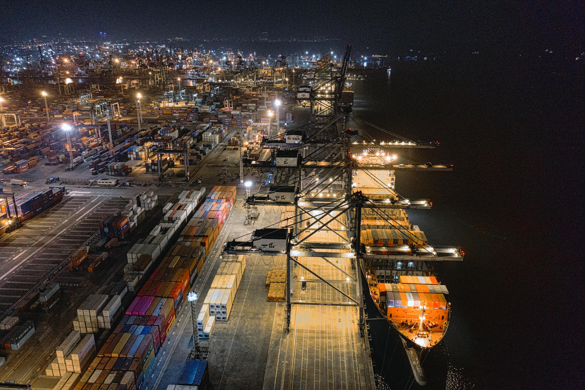 Widok na port o zmroku. W oświetlonym porcie stoi statek towarowy oraz setki kontenerów.